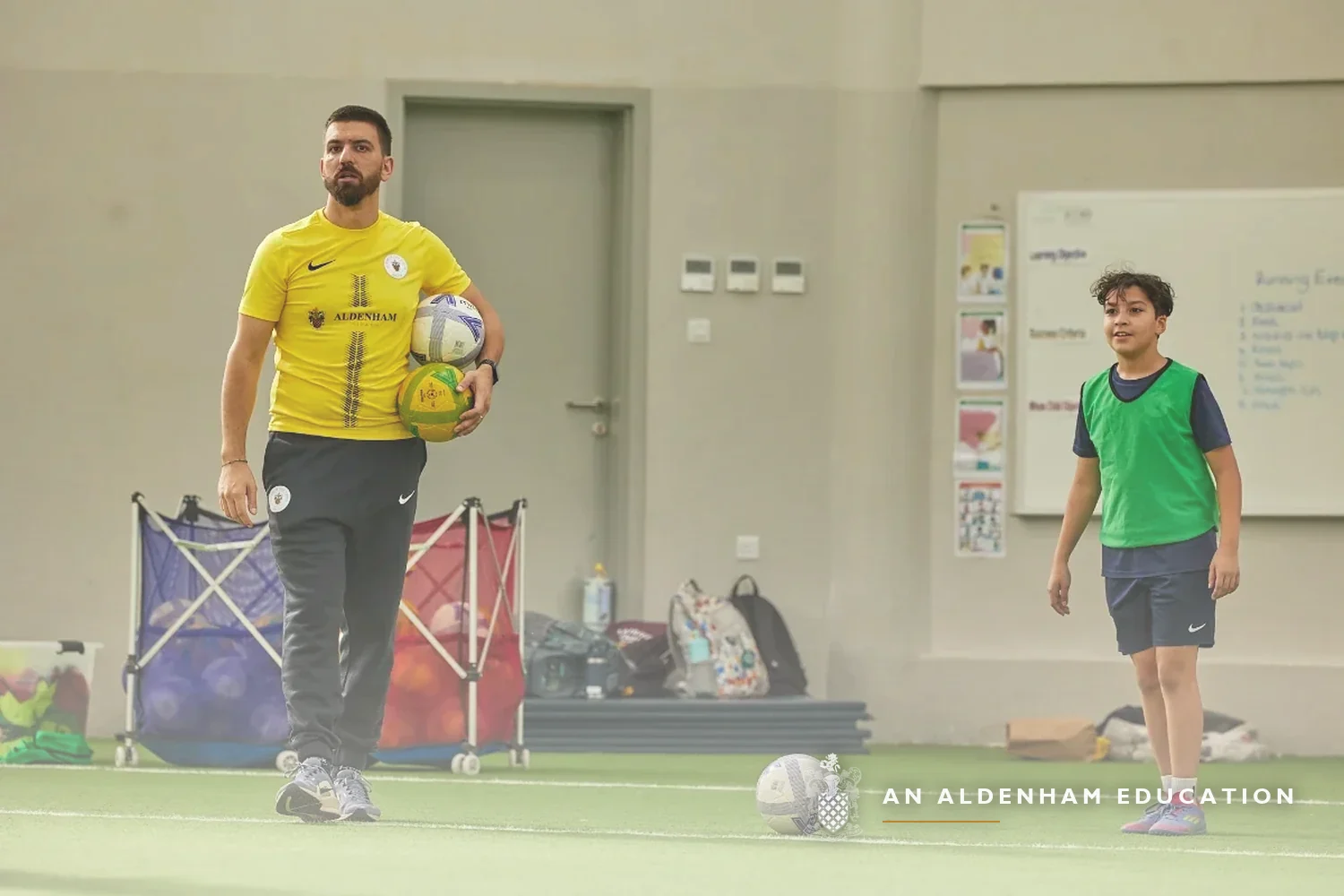 A coach in yellow Academy kit holding footballs, leading a training session with a young player on the indoor pitch