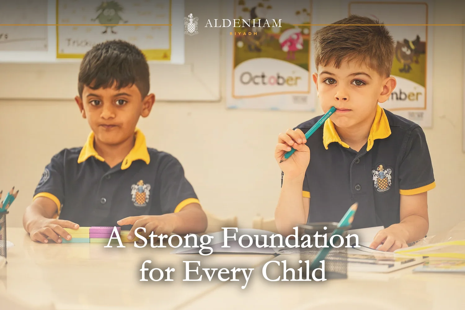 Two young pupils in navy and gold Aldenham School Riyadh uniform at their classroom desk, one holding a pencil, with phonics posters behind